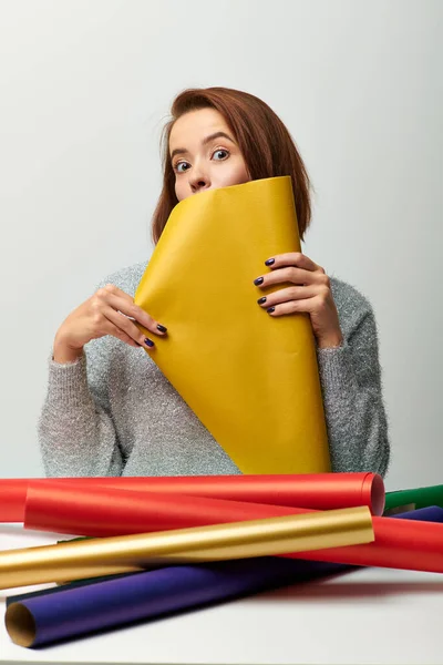 Temporada de alegría, mujer joven en suéter sosteniendo papel de embalaje amarillo sobre fondo gris, tiempo de Navidad - foto de stock