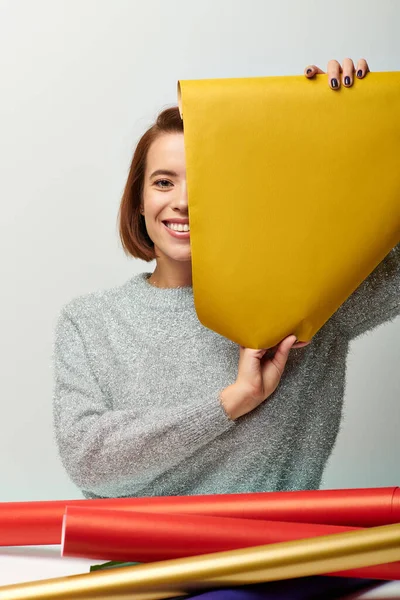 Сезон радості, весела жінка в светрі тримає жовтий подарунковий папір на сірому тлі, Різдвяний час — стокове фото