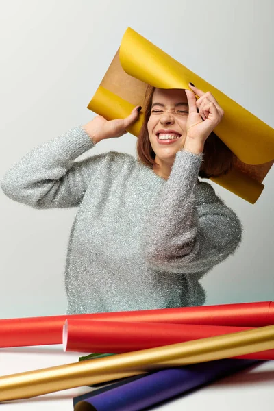 Cheerful woman in sweater wrapping herself in yellow gift paper on grey backdrop, Christmas joy — Stock Photo