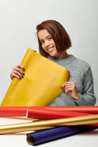 Smiling woman in cozy sweater holding yellow gift paper on grey backdrop, Merry Christmas concept — Stock Photo
