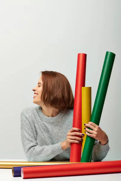 Joyous woman in cozy sweater holding different colors gift paper on grey backdrop, Merry Christmas — Stock Photo