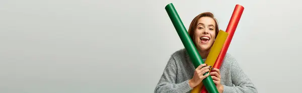 Excited woman in winter sweater holding colorful wrapping paper on grey background, Christmas banner — Stock Photo