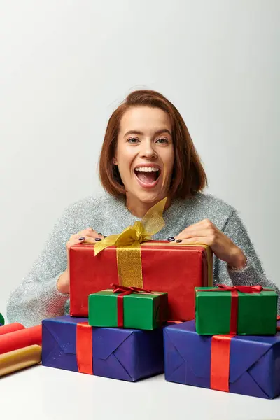 Donna eccitata in maglione invernale vicino regalo avvolto e carta regalo colorata su grigio, Natale — Foto stock