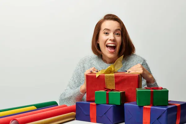 Femme étonnée en pull d'hiver près de cadeau enveloppé et papier cadeau coloré sur gris, Noël — Photo de stock