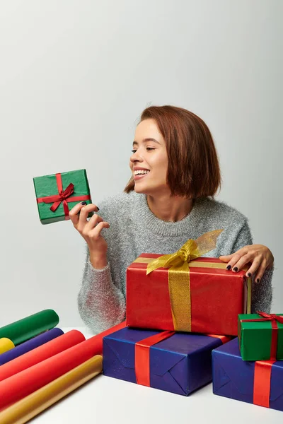 Femme heureuse en pull d'hiver regardant enveloppé cadeau de Noël près de papier cadeau coloré sur gris — Photo de stock