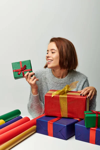 Felice donna in maglione invernale guardando avvolto regalo di Natale vicino carta regalo colorato su grigio — Foto stock