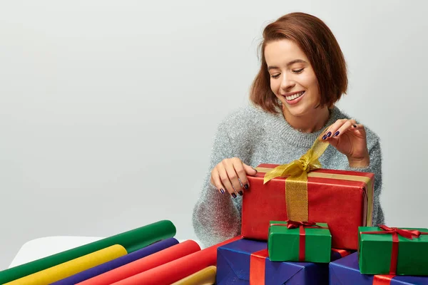 Femme heureuse en pull d'hiver tirant ruban sur enveloppé cadeau de Noël sur fond gris — Photo de stock