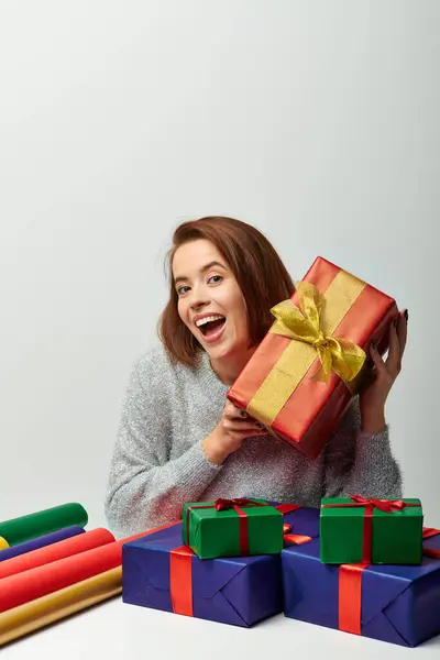 Donna entusiasta in maglione invernale in possesso di regalo di Natale rosso vicino carta regalo su sfondo grigio — Foto stock