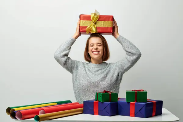 Cheerful woman in winter sweater holding Christmas present above head near gift paper on grey — Stock Photo