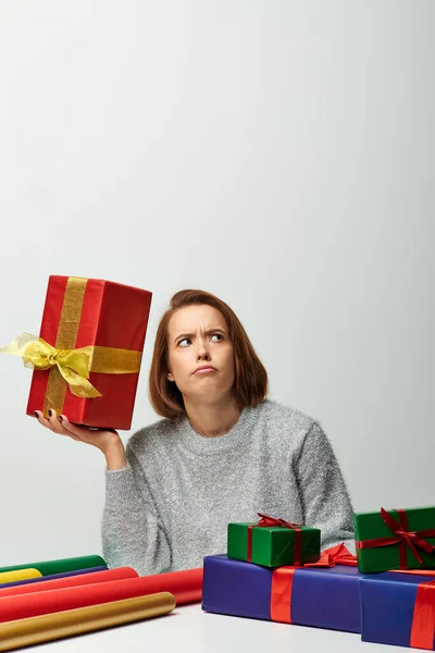 Любопытная женщина в трикотажном свитере держит рождественский подарок рядом с красочной подарочной бумагой на сером фоне — стоковое фото
