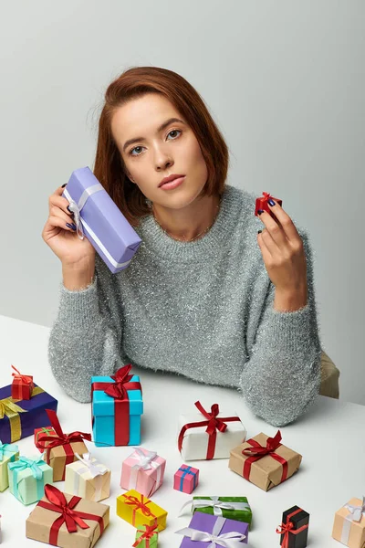Брюнетка в уютном свитере с маленьким рождественским подарком рядом с красочными завернутыми подарками на сером — стоковое фото
