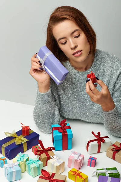 Morena mujer en suéter acogedor mirando regalo de Navidad cerca de coloridos regalos envueltos en gris - foto de stock