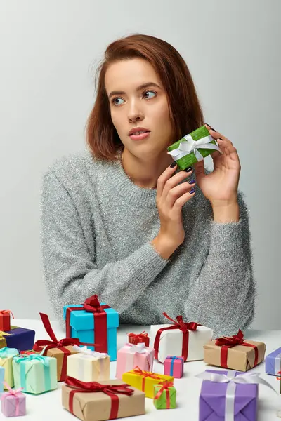 Beautiful woman in cozy sweater holding tiny Christmas present near colorful wrapped gifts on grey — Stock Photo