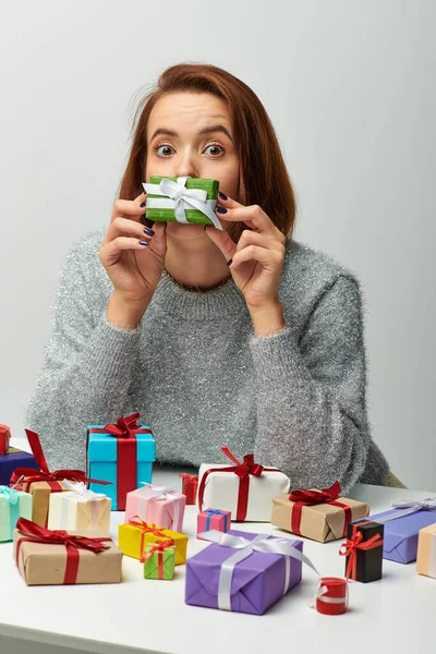 Mujer divertida en suéter sosteniendo pequeño regalo de Navidad cerca de la cara y coloridos regalos envueltos en gris - foto de stock