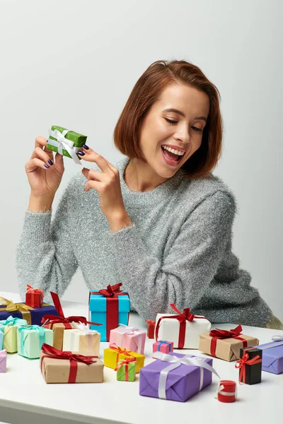 Mujer emocionada en suéter acogedor celebración pequeño regalo de Navidad cerca de coloridos regalos envueltos en gris - foto de stock