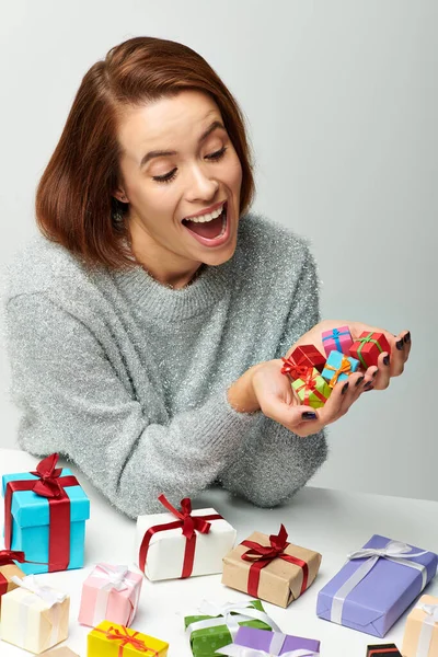 Взволнованная женщина в уютном свитере смотрит на кучу крошечных рождественских подарков рядом с красочными завернутыми подарками — стоковое фото