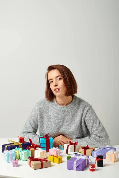 Attraente donna in maglione seduta tra mazzi di regali di Natale sul tavolo, sfondo grigio — Foto stock