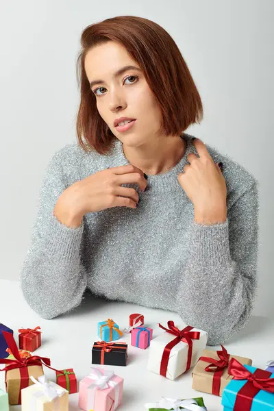 Bella donna in maglione invernale seduta tra mazzi di regali di Natale sul tavolo, sfondo grigio — Foto stock