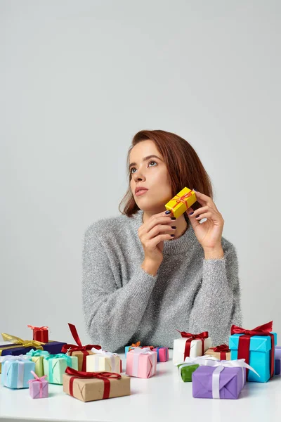 Pretty woman in sweater fantasizing and holding Christmas gift near bunch of presents on table — Stock Photo