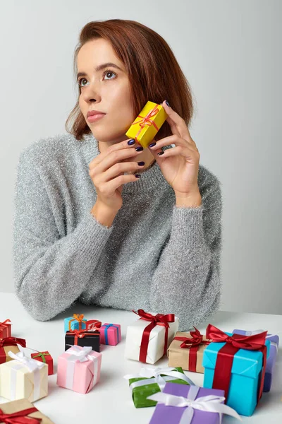 Привлекательная женщина в свитере фантазирует и держит рождественский подарок рядом с кучей подарков на столе — стоковое фото