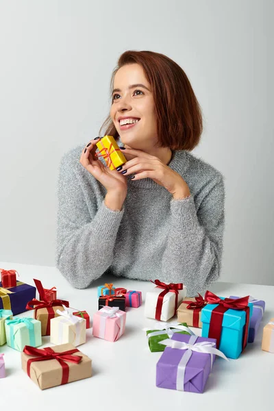 Donna allegra in maglione fantasticando e tenendo regalo di Natale vicino mazzo di regali sul tavolo — Foto stock