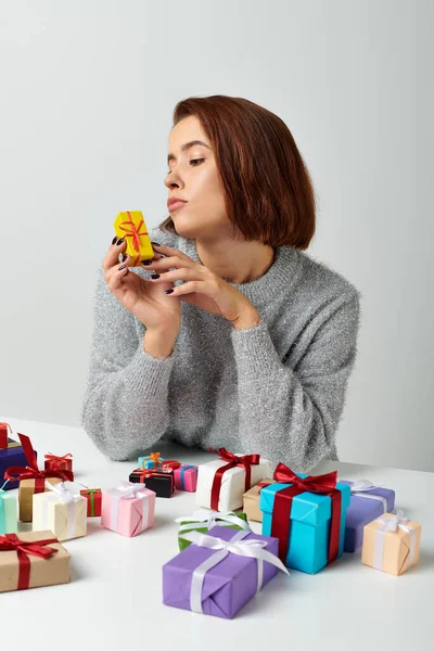 Giovane donna in maglione guardando piccolo regalo di Natale vicino mazzo di regali sul tavolo, sfondo grigio — Foto stock