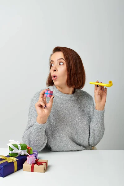 Shocked woman in sweater holding tiny Christmas gift and yellow party horn on grey backdrop — Stock Photo