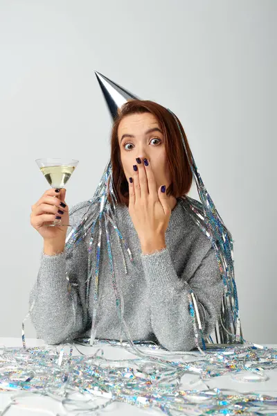 Young woman in party cap with tinsel on head holding glass of champagne and covering face with hand — Stock Photo