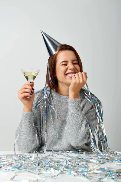 Thrilled woman in party cap with tinsel on head holding glass of champagne on grey, Happy New Year — Stock Photo