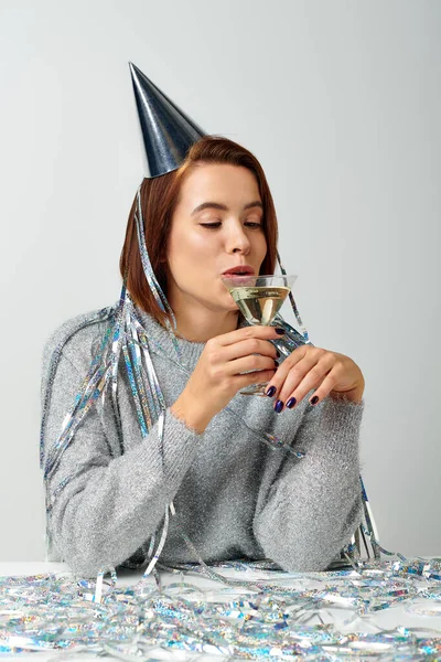 Jovem mulher em boné de festa com ouropel na cabeça bebendo champanhe em pano de fundo cinza, Feliz Ano Novo — Fotografia de Stock
