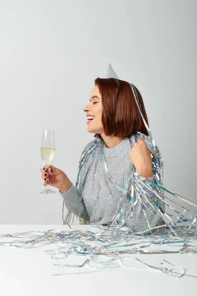 Joyful woman in party cap and tinsel on head holding glass of champagne while celebrating New year — Stock Photo