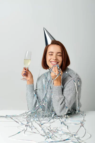 Happy woman in party cap and tinsel on head holding champagne glass while making a wish on New year — Stock Photo