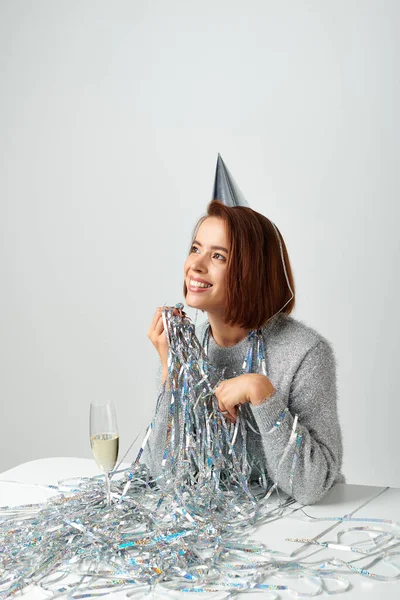 Satisfied woman in party cap and tinsel smiling near champagne glass while celebrating New year — Stock Photo