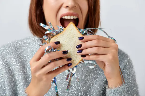 Close up of cropped woman biting sandwich with tinsel on grey backdrop, Happy New Year concept — Stock Photo