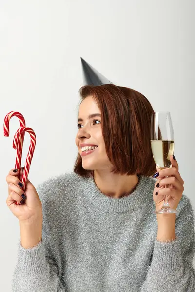 Щаслива жінка у вечірній кепці, що тримає келих шампанського та цукерки на сірому, концепція веселого Різдва — стокове фото