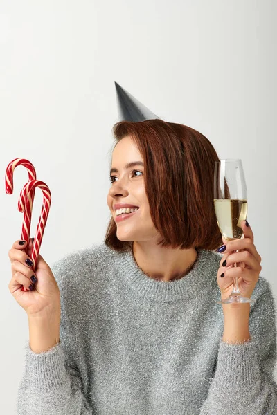 Happy woman in party cap holding champagne glass and candy canes on grey, Merry Christmas concept — Stock Photo