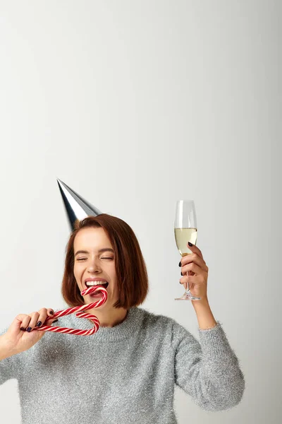 Young woman in party cap holding champagne glass and biting candy cane on grey, Merry Christmas — Stock Photo