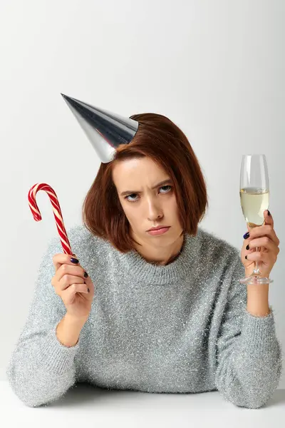 Незадоволена жінка у вечірній кепці тримає келих шампанського та цукеркову тростину на сірому, з Різдвом — стокове фото