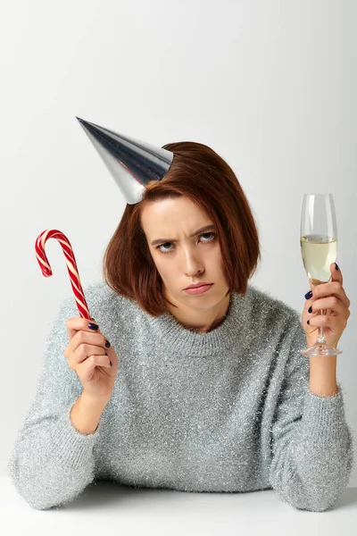 Displeased woman in party cap holding champagne glass and candy cane on grey, Merry Christmas — Stock Photo
