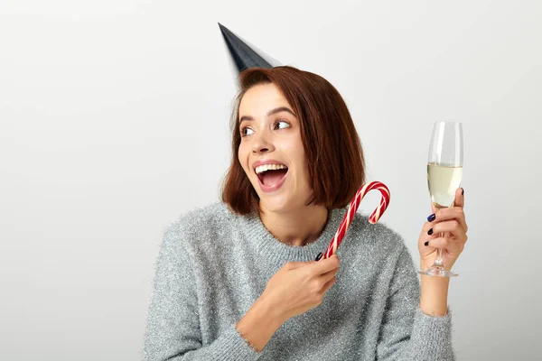 Excited woman in party cap holding champagne glass and candy cane on grey, Merry Christmas — Stock Photo