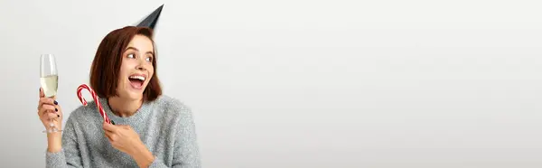 Excited woman in party cap holding champagne glass and candy cane on grey, Merry Christmas, banner — Stock Photo