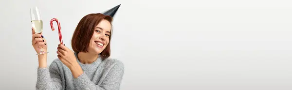 Positive woman in party cap holding champagne glass and candy cane on grey, Merry Christmas, banner — Stock Photo