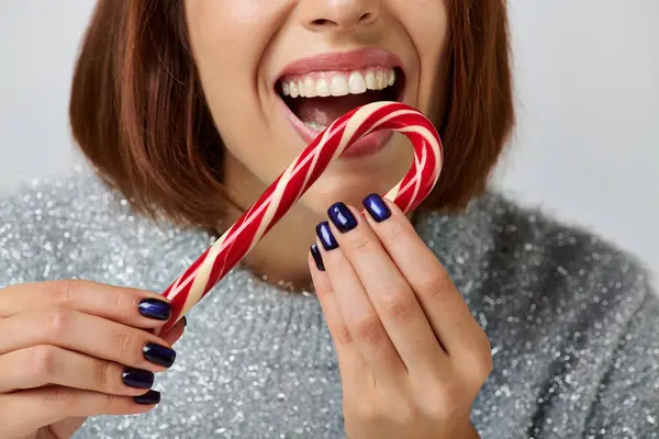 Cropped cheerful woman in grey sweater biting tasty candy cane on grey, Merry Christmas concept — Stock Photo
