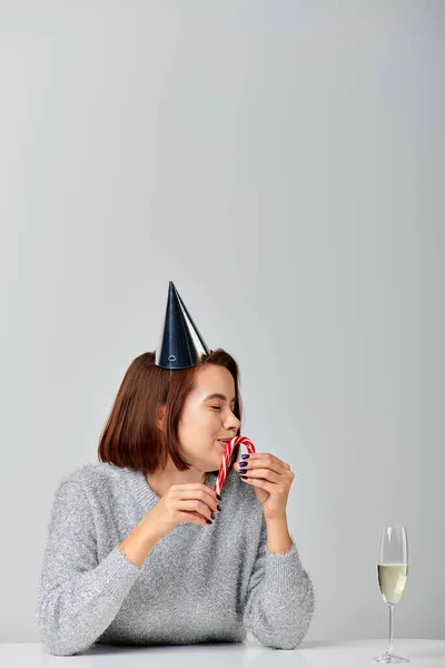 Happy woman in party cap kissing candy cane near champagne glass on grey backdrop, Merry Christmas — Stock Photo