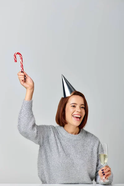 Щаслива жінка у вечірній кепці, що тримає цукеркову тростину та келих шампанського на сірому фоні, з Різдвом — стокове фото
