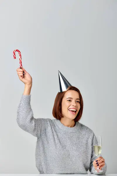 Thrilled woman in party cap holding candy cane and champagne glass on grey backdrop, Merry Christmas — Stock Photo
