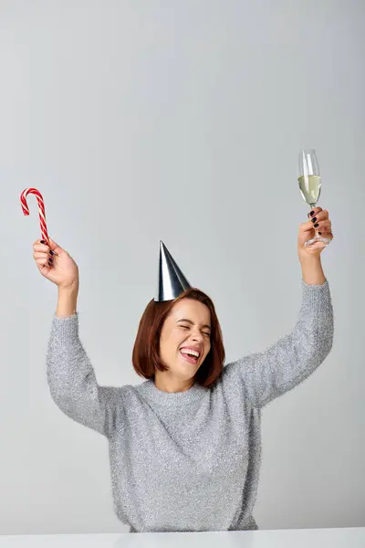 Збуджена жінка у вечірній кепці, що тримає цукеркову тростину та келих шампанського на сірому фоні, щасливий Новий рік — стокове фото