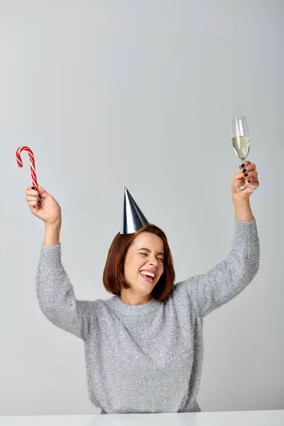 Excited woman in party cap holding candy cane and champagne glass on grey backdrop, Happy New Year — Stock Photo