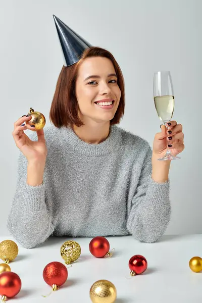 Happy woman in party cap holding champagne glass and shiny bauble on grey, Merry Christmas concept — Stock Photo