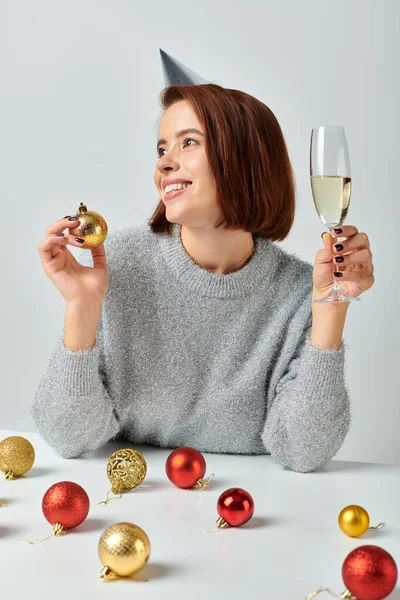 Allegra donna in berretto partito tenendo bicchiere di champagne e bauble su grigio, Buon Natale concetto — Foto stock